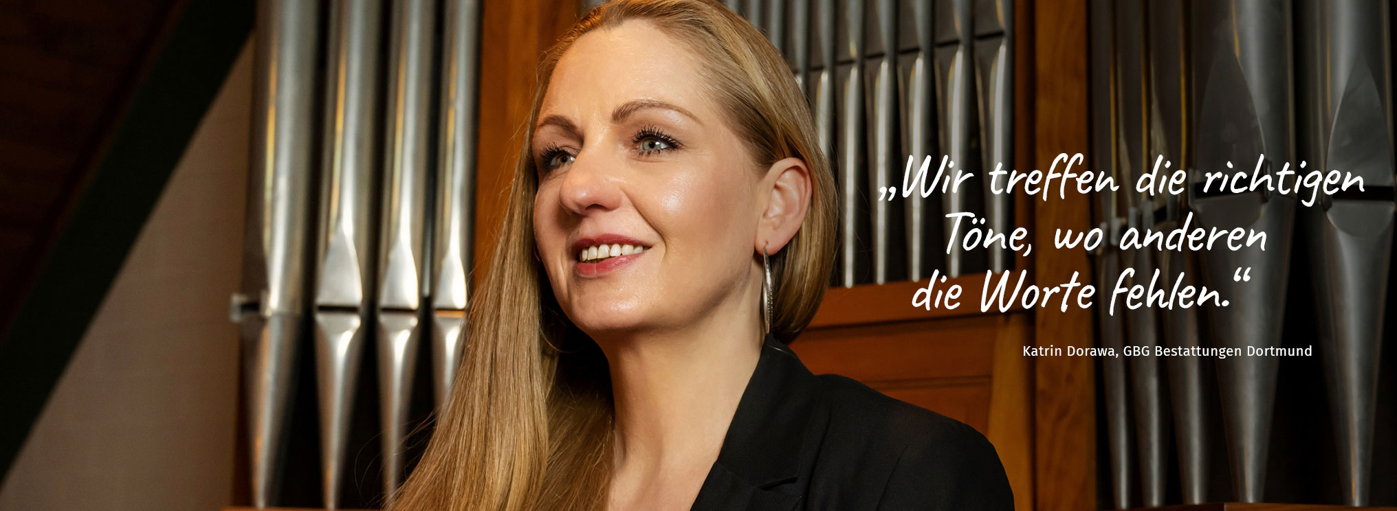 Katrin Dorawa von GBG Bestattungen Dortmund vor einer Orgel, lächelnd und in schwarzer Kleidung. Der Text auf dem Bild lautet: „Wir treffen die richtigen Töne, wo anderen die Worte fehlen.“