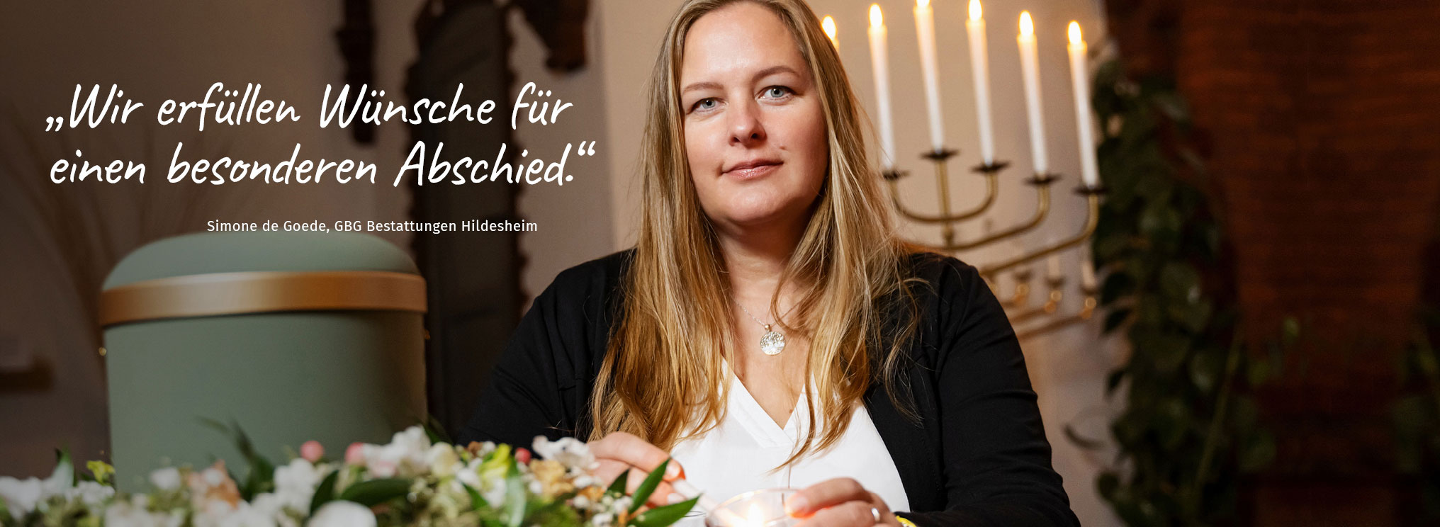 Mitarbeiterin von GBG Bestattungen Hildesheim entzündet eine Kerze neben einer Urne, mit Blumenschmuck und Kerzenleuchter im Hintergrund. Der Text auf dem Bild lautet: „Wir erfüllen Wünsche für einen besonderen Abschied.“ – Simone de Goede, GBG Bestattungen Hildesheim.